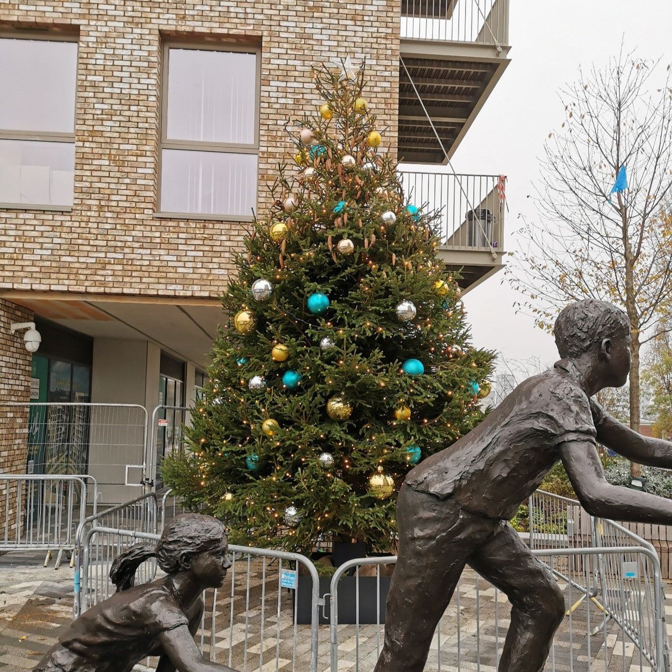 Bronze statues of children playing in front of a decorated Christmas tree outside a building.
